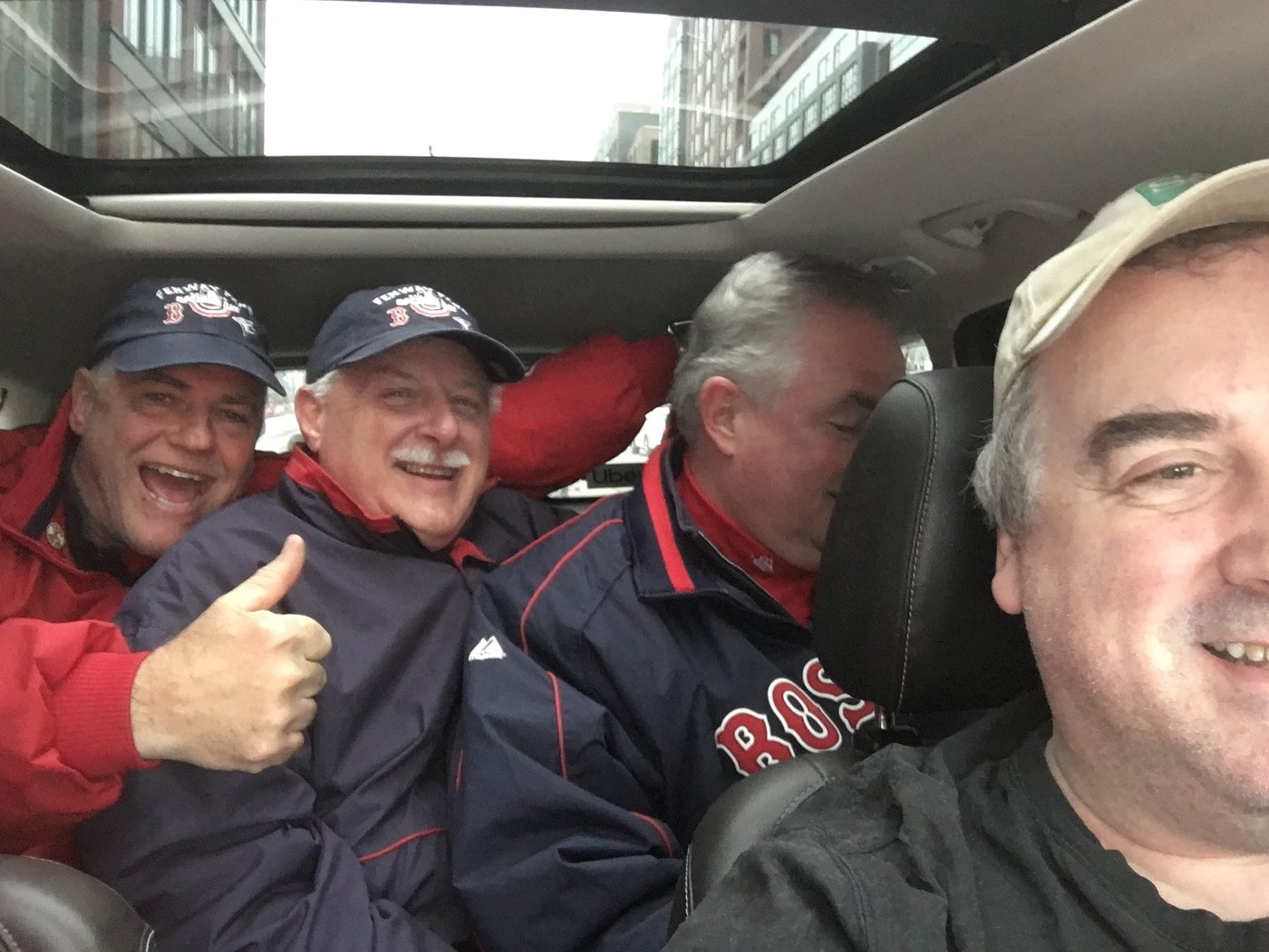 Four men in a car on a Boston street, three riders in Fenway Park caps and Red Sox jackets, one giving a thumbs up, with the driver at the wheel.